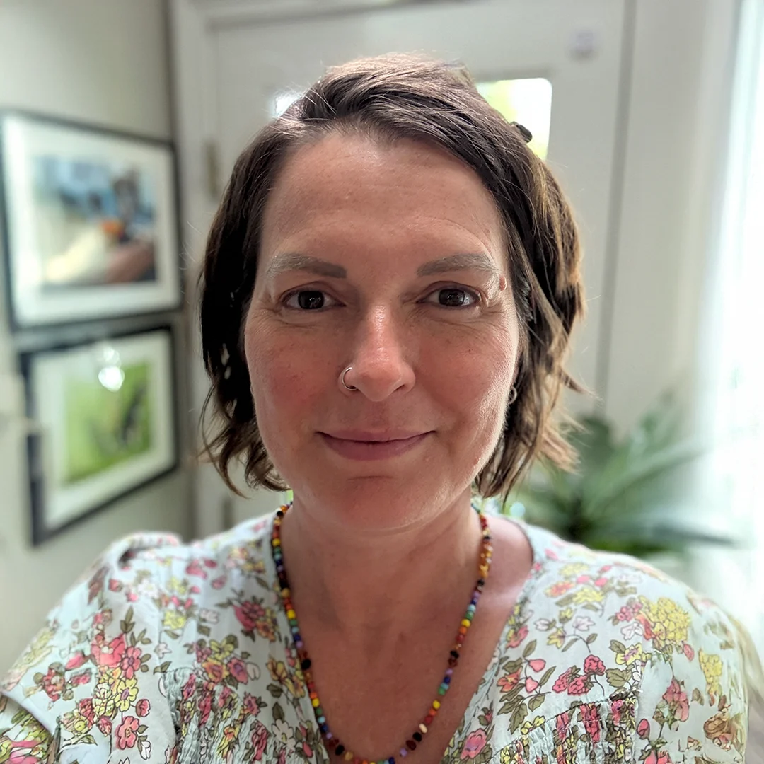 Dr. Soco smiling warmly in a floral top, wearing a colorful beaded necklace, with soft natural light in the background.